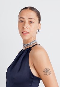 Top de culoare navy, cu gât halter, cu o textură netedă, având un decolteu plisat și un colier din strasuri argintii. Brațul prezintă o tatuaj floral.