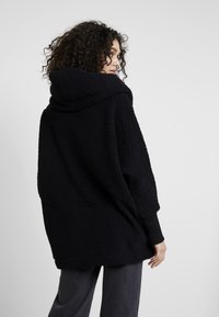 Hoodie en polaire noir texturé avec une grande capuche, coupe ample, et poignets côtelés, présenté de côté.