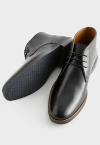 Botas de tobillo de cuero negro con textura suave, punta redondeada, diseño de cordones, costuras en contraste de color marrón y suela de goma texturizada.