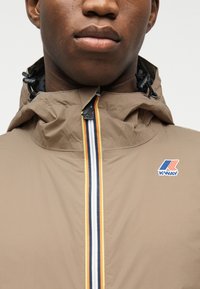 Κοντινή λήψη ενός ατόμου που φοράει ένα μπεζ μπουφάν K-Way με ρίγα φερμουάρ και ρυθμιζόμενα κορδόνια κουκούλας.