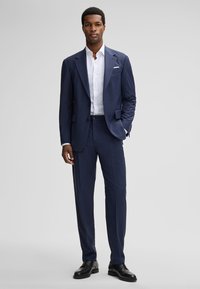 Man staat zelfverzekerd in een marineblauw pak, een witte overhemd, zwarte dresschoenen en een witte pochet tegen een grijze achtergrond.