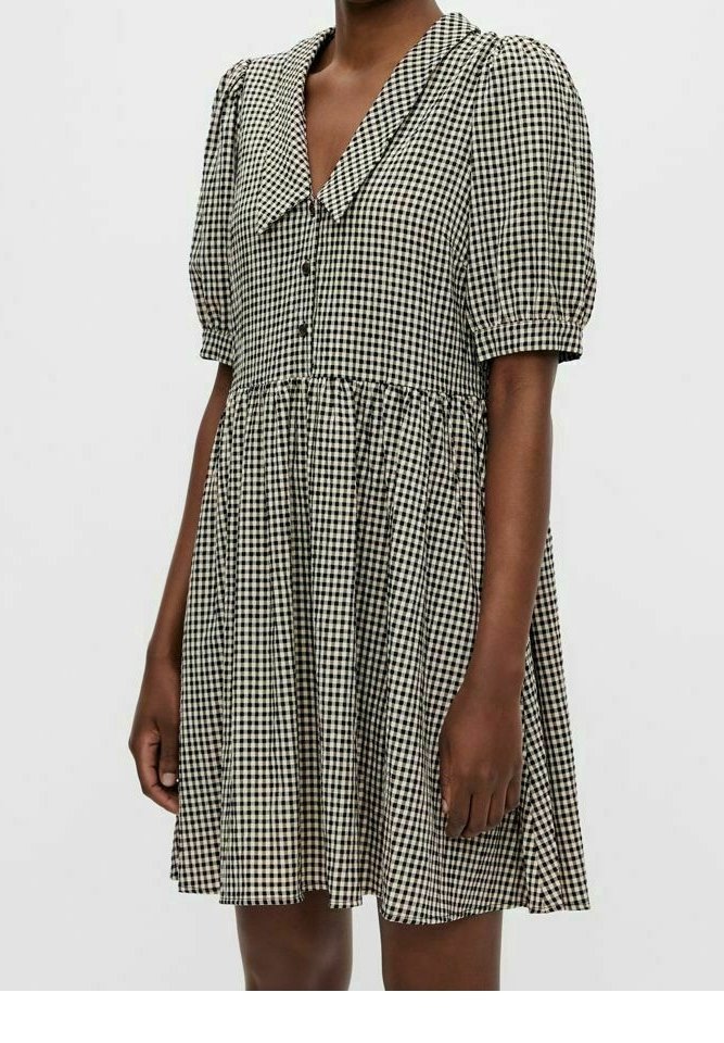 Robe vichy noire et blanche à manches bouffantes, avec des boutons sur le devant et un large col, portée par une personne debout sur un fond uni.