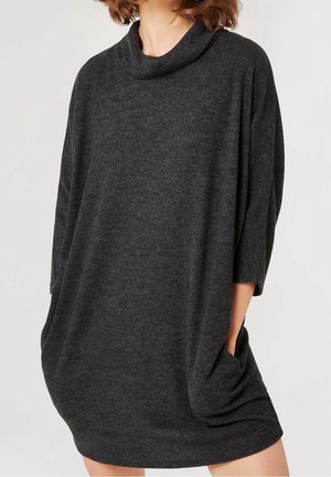 Kobieta ubrana w luźną, ciemnoszarą dzianinową sukienkę z trzy czwarte rękawami i kołnierzem typu komin, z rękami wsuniętymi do bocznych kieszeni.