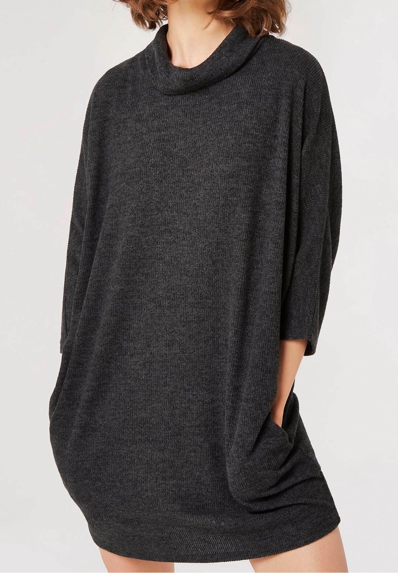 Femme portant une robe pull ample en maille gris foncé avec des manches trois quarts et un col bénitier, mains glissées dans les poches latérales.