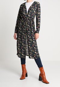 Blommig lång cardigan i svart med gula, vita och lila mönster, med långa ärmar och knytmidja, som kombineras med orange klackstövlar.