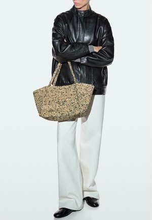 Osoba ve černé kožené bundě a bílých širokých kalhotách drží velkou tkanou tašku s krátkými uchy.