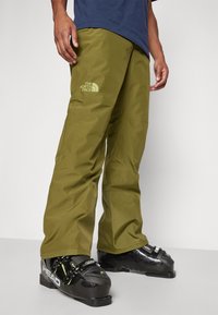 Green ski pants with a relaxed fit, featuring The North Face logo on the thigh and paired with black ski boots, showcasing durable material.