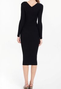 Svart ribbad stickad klänning med lång ärm, åtsittande form och V-ringning bak. Knälång, parat med beiga sandaler med remmar.