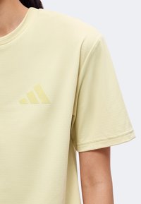 Vue latérale d'une personne portant une chemise jaune clair à manches courtes avec un logo subtil à trois bandes sur la poitrine.