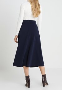 Jupe midi marine en tissu lisse, dotée d'un design plissé et d'une fermeture éclair cachée à l'arrière. Portée avec des bottines marron.