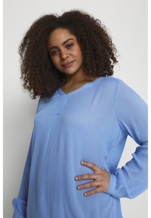 Lichtblauw languit shirt met V-hals en een zak aan de voorkant, met een gestructureerde stof en een losse pasvorm. Model poseert met de hand op de heup.