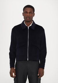 Navy corduroy bomber jacket with a collared neck, front zip, and two large patch pockets. Textured fabric and cropped design.