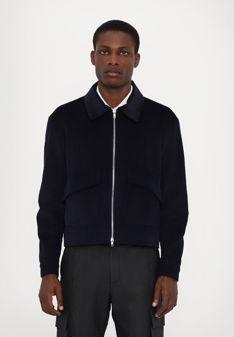 Navy corduroy bomber jacket with a collared neck, front zip, and two large patch pockets. Textured fabric and cropped design.