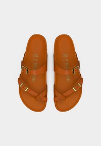 Πορτοκαλί σανδάλια κατασκευασμένα από συνθετικό υλικό με δύο φαρδιές λουρίδες και δύο ρυθμιζόμενες μεταλλικές αγκράφες. Λείο, επίπεδο πέλμα με ανάγλυφο λογότυπο.