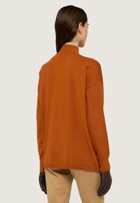 Rostfarbener Pullover mit langen Ärmeln und Stehkragen, weicher Textur und lässiger Passform. Getragen mit braunen Lederhandschuhen, kombiniert mit beigen Hosen.