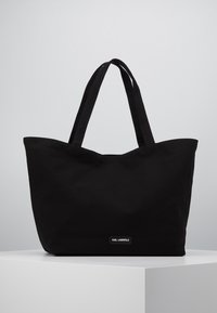Black tote bag made of durable fabric, featuring two sturdy handles and a minimalist design. A small logo tag is attached at the bottom.