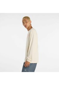 Langärmliges, cremefarbenes T-Shirt mit grafischen Details in Rot und Grün, präsentiert von einem Modell, das graue Shorts trägt, im Profil.