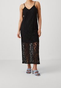 Black lace dress with a V-neck, adjustable straps, and a sheer lower skirt. Paired with light blue heeled shoes.