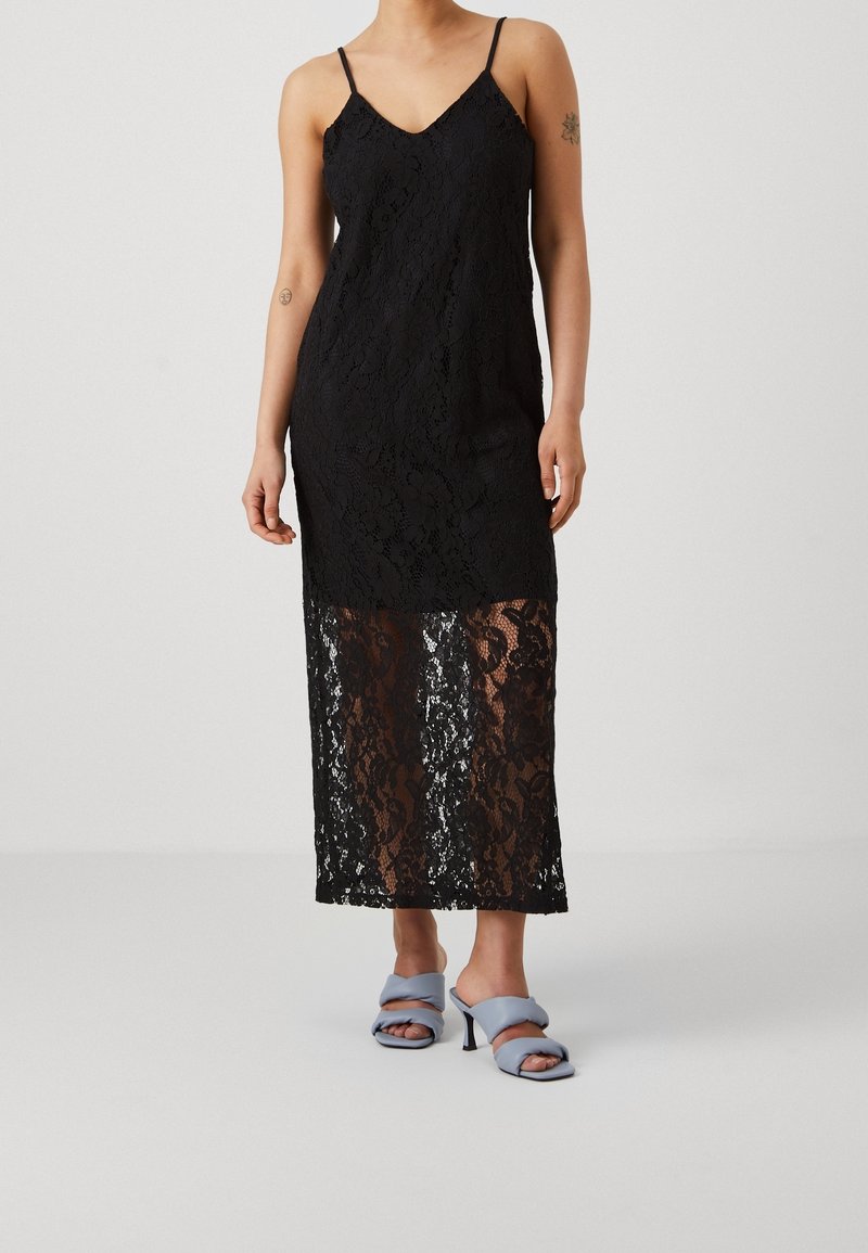 Black lace dress with a V-neck, adjustable straps, and a sheer lower skirt. Paired with light blue heeled shoes.