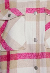 Camicia a quadri con una miscela di rosa, beige e bianco. Presenta due tasche sul petto, pattine arrotondate per le tasche e bottoni a pressione metallici.