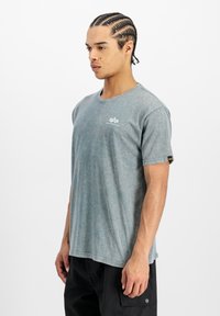 Noor mees, kellel on punutud juuksed, kannab pleekinud sinist Alpha Industries t-särki ja musti taskupükse, seistes külgsuunas valge tausta vastu.