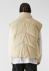 Beige padded vest with a high collar, featuring chevron stitching and a slightly rounded hem. Material appears soft and smooth.