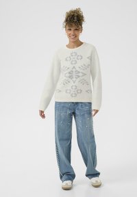Pull en tricot blanc avec un motif géométrique argenté, associé à un jean large bleu clair orné d'accents en perles et des baskets blanches.