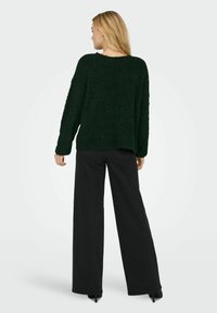 Grüner, flauschiger Pullover mit rundem Ausschnitt, langen Ärmeln und lockerer Passform, kombiniert mit hochtaillierten schwarzen weit geschnittenen Hosen und Absätzen.