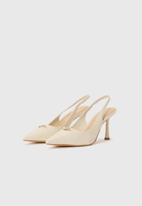 Tacchi slingback beige con punta affusolata, un piccolo accent oro sul davanti e un tacco sottile. Texture liscia e design minimalista.