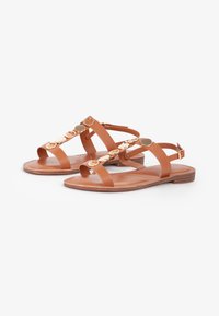 Sandalias de cuero marrón claro con suelas planas, que presentan múltiples correas horizontales y acentos metálicos circulares a lo largo del centro.