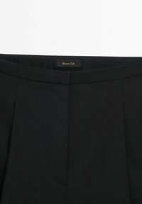 Mustat räätälöidyt housut, joissa on sileä pinta, litteä edusta, laskokset vyötäröllä ja logotunniste. Kangas näyttää kevyeltä ja juhlavalta.