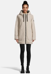 Beige gefütterter Mantel mit Kapuze, Frontreißverschluss, schwarzen Kordeln und Logo-Akzent am Ärmel, kombiniert mit schwarzen Leggings und Stiefeln.