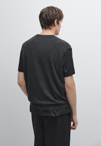 Man staat met zijn rug naar de camera gekeerd, draagt een donkergrijs mouwloos vest over een zwart T-shirt met korte mouwen en zwarte broek.