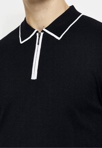 Czarna dzianinowa koszulka polo z białym wykończeniem przy kołnierzu i półzamkiem noszona przez osobę, przycięta na szyi i klatce piersiowej.