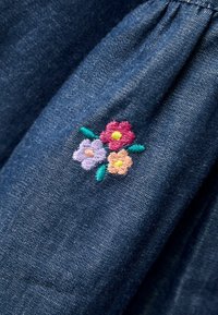 Denimová látka v tmavomodrej farbe s nariaseným detailom; obsahuje vyšívané kvety v ružovej, fialovej a žltej farbe s zelenými listami.