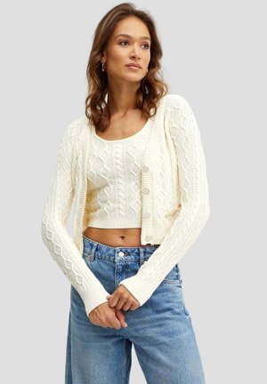 Cardigan bianco a maglia con intricati motivi a diamante, design cropped, polsini a coste e chiusura con bottoni, abbinato a jeans a vita alta.