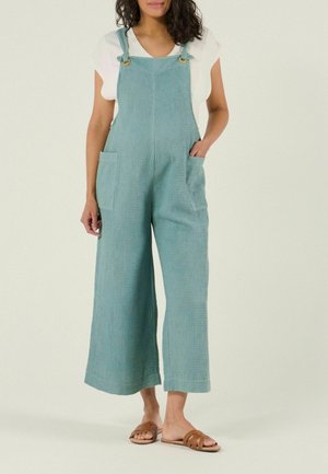 Femme portant une salopette large en tissu gaufré couleur sarcelle avec poches et des sandales marron à enfiler sur un fond clair.