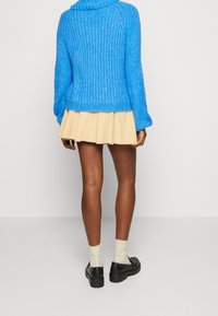 Personne portant un pull tricoté épais bleu vif, une mini-jupe plissée beige, des chaussettes crème et des mocassins à plateforme noirs, debout de dos.
