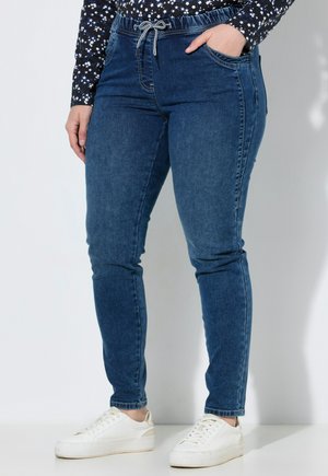 Person wearing blue denim jogger jeans with a drawstring waist and white sneakers, standing with one hand in pocket.