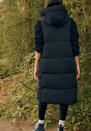 Gilet long noir rembourré avec une capuche, présentant un tissu texturé sur les manches. Porté sur des leggings noirs et des chaussures montantes avec des accents blancs.