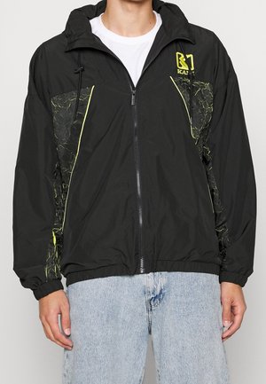 Person trägt eine schwarze Jacke mit Reißverschluss, neongelben Akzenten und Kapuze, kombiniert mit hellblauen Jeans.