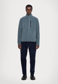 Veste en polaire bleu clair avec col montant, fermeture éclair et poche poitrine noire, associée à un pantalon bleu foncé et des chaussures noires.