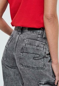 Une personne portant un jean en denim noir délavé à taille haute avec de grandes poches arrière et une chemise rouge rentrée, montrant des détails sur le côté et l'arrière.
