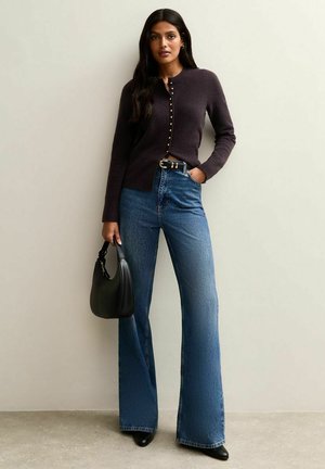 Donkerpaarse cardigan met knopen, gecombineerd met high-waisted flared spijkerbroeken. Accessoires zijn een zwarte tas en een riem. Soepele texturen.