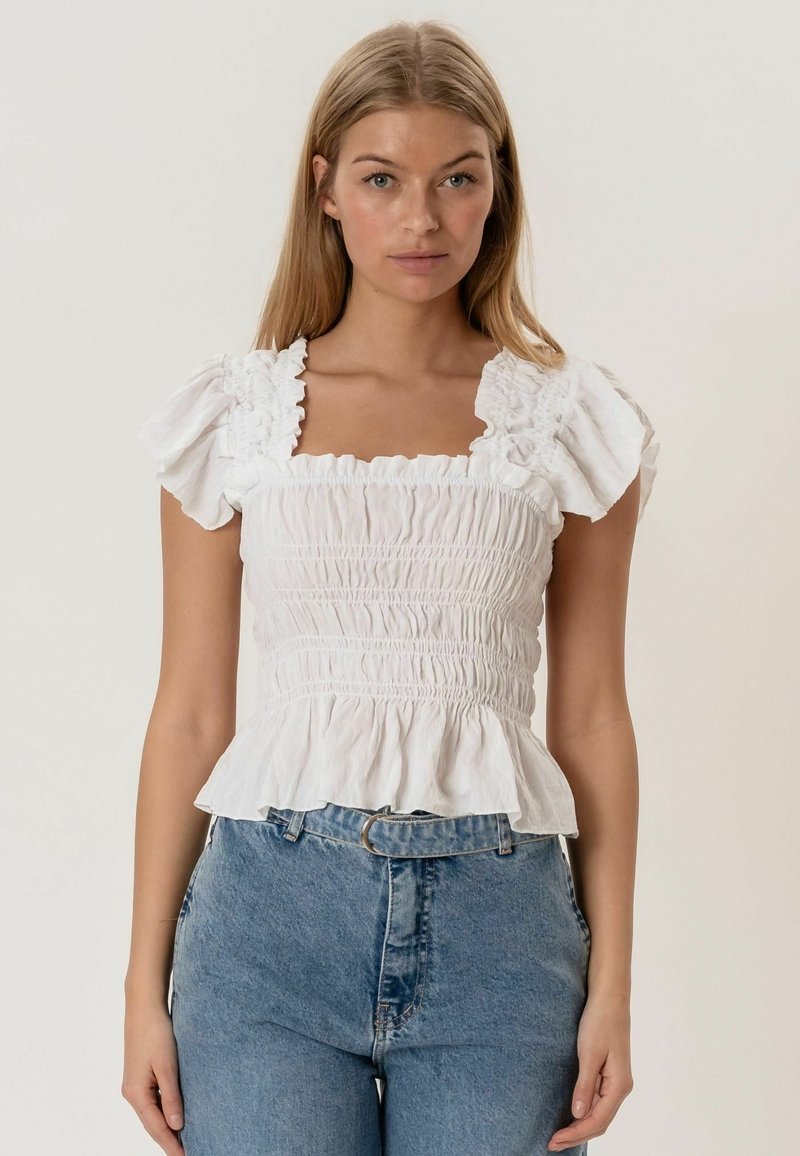 Femme blonde portant un chemisier blanc à encolure carrée froncée avec des manches à volants et un jean bleu à taille haute, posant devant un fond uni.