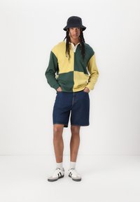 Sudadera de bloques de color en amarillo y verde con un cuello blanco, combinada con shorts de mezclilla azules, zapatillas blancas y un sombrero de cubo negro.