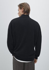 Zwarte gebreide trui met een hoge kraag, ontspannen pasvorm en geribde zoom. Het materiaal lijkt zacht en textuur te hebben, geschikt voor lagen.