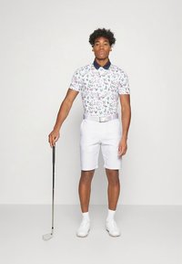 Golfpolo med korta ärmar och ett blommigt mönster i rosa och grönt, kombinerad med vita shorts och ett ljust bälte, håller i en golfklubba.