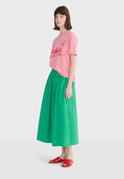 Młoda kobieta stoi na tle jasnego, jednolitego tła, ubrana w różowy t-shirt z grafiką, długą zieloną spódnicę i czerwone sandały na otwartym palcu.