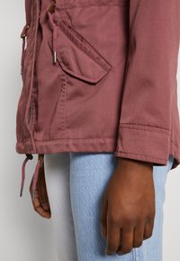 Růžová látková bunda se staženým pasem, dvěma šikmými kapsami a šňůrkami na lemu, doplněná světle modrými džínami.